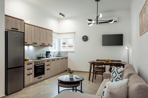 A modern kitchen with a refrigerator, oven, and coffee maker, a dining area with a table and chairs, and a living room with a sofa and a TV mounted on the wall. - Marilianas Apartment (Platanias)