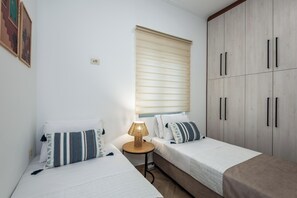 A bedroom with two single beds, a bedside table with a lamp, a wardrobe, and a window with blinds. - Marilianas Apartment (Platanias)