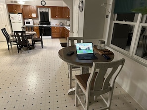 A kitchen with wooden cabinets, a dining table with chairs, a laptop on the table, and a window with a view.