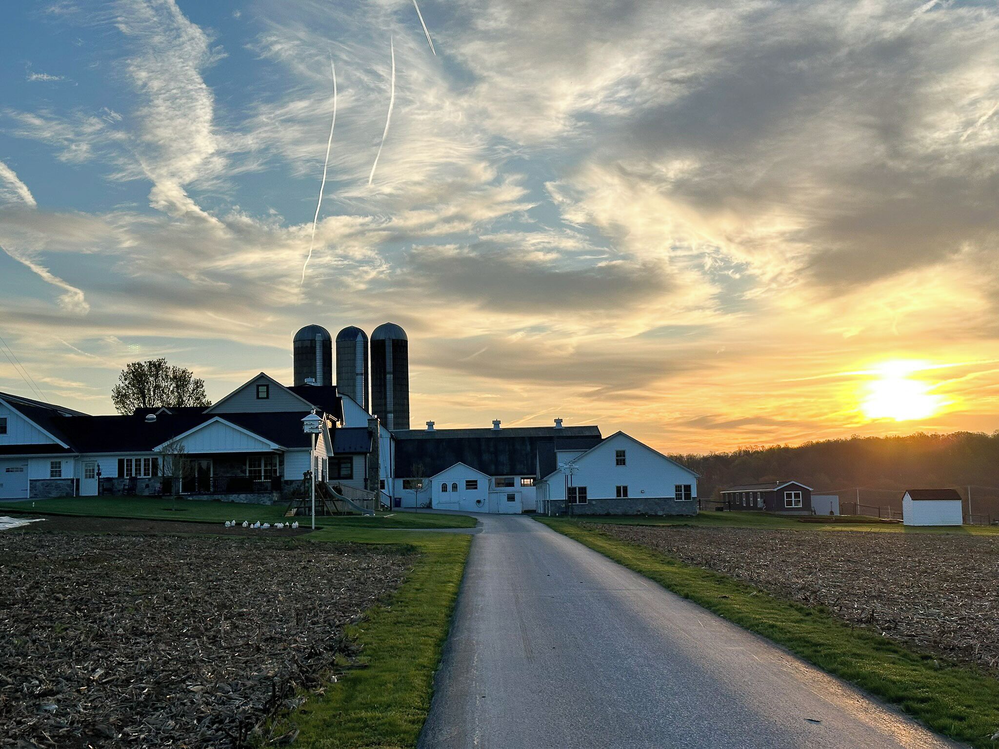 A rural landscape with a farmhouse, silo buildings, and a clear sky at sunset.