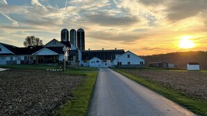 A rural landscape with a farmhouse, silo buildings, and a clear sky at sunset.