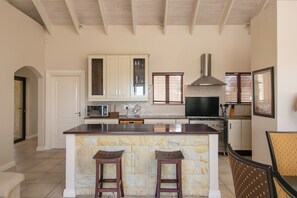 Une cuisine dotée d’un îlot central, de deux tabourets de bar, d’un four à micro-ondes, d’une cuisinière et d’une grande fenêtre avec des stores.