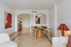 A modern dining area with a round table and chairs, a white sofa, and a lamp. - Elegant 2BR in Los Arqueros Golf & Country Club (Benahavís)