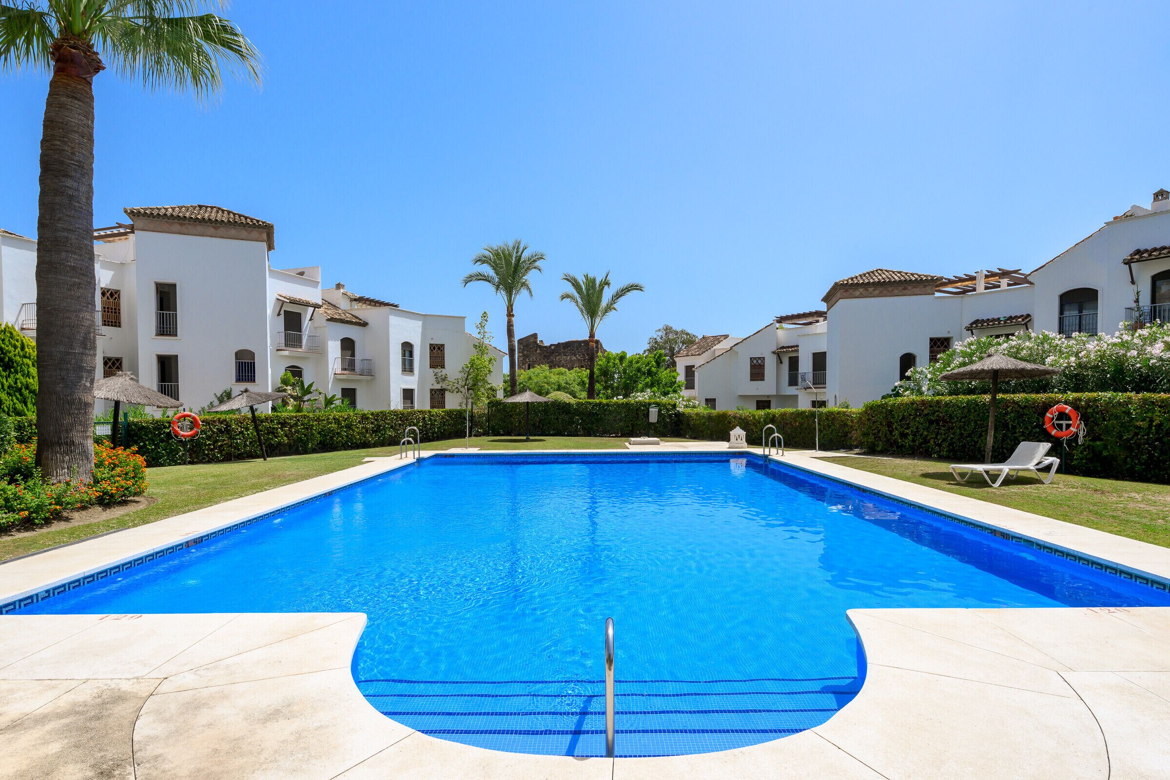 A clear blue swimming pool with a diving board, surrounded by white buildings with balconies and palm trees.