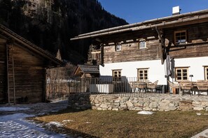 Un edificio rústico de madera con base de piedra, un balcón con mobiliario exterior y montañas al fondo.