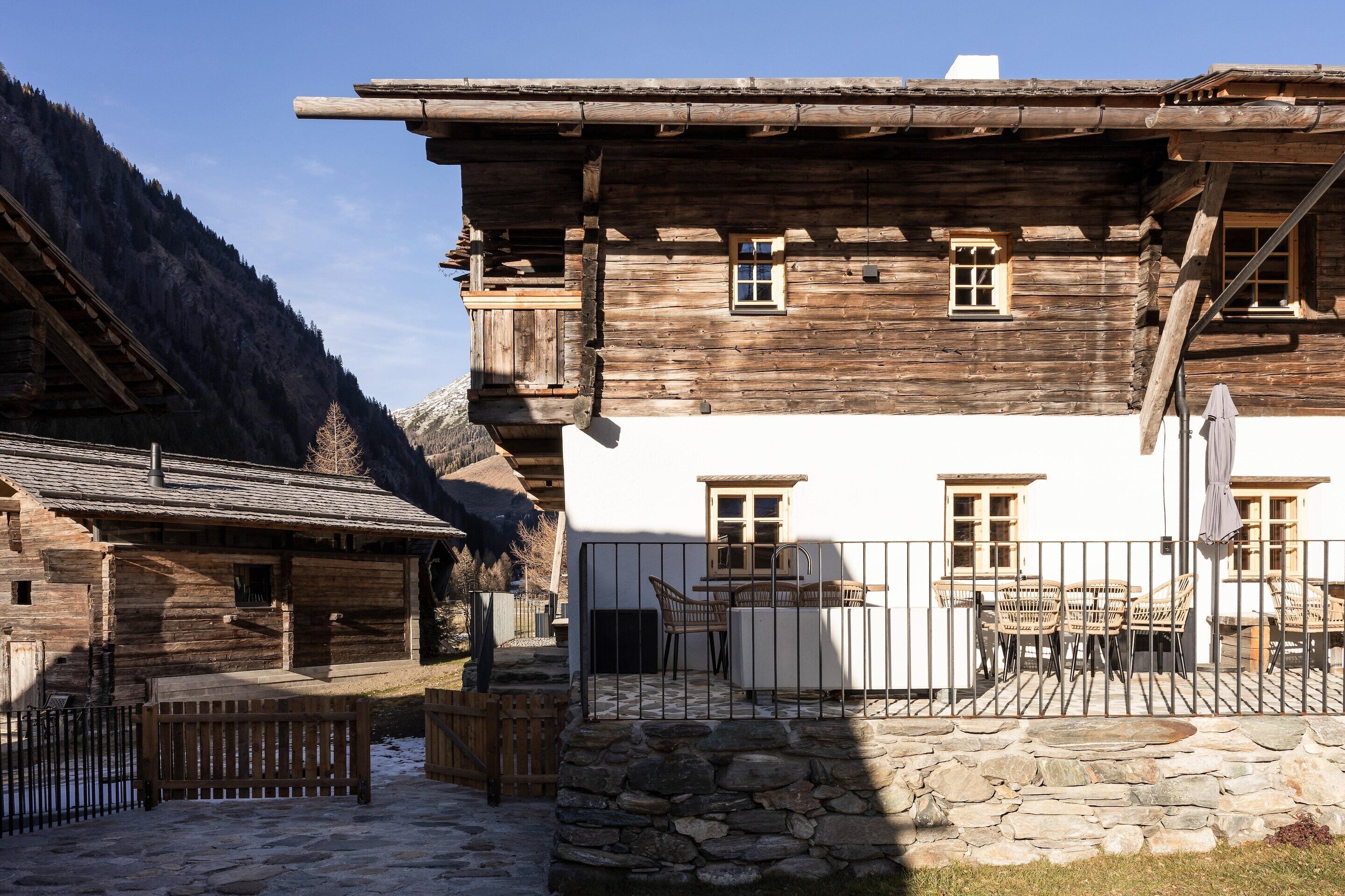 Un edificio tradicional de madera con base de piedra, un balcón con mobiliario exterior y montañas al fondo.