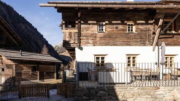 Un edificio tradicional de madera con base de piedra, un balcón con mobiliario exterior y montañas al fondo.