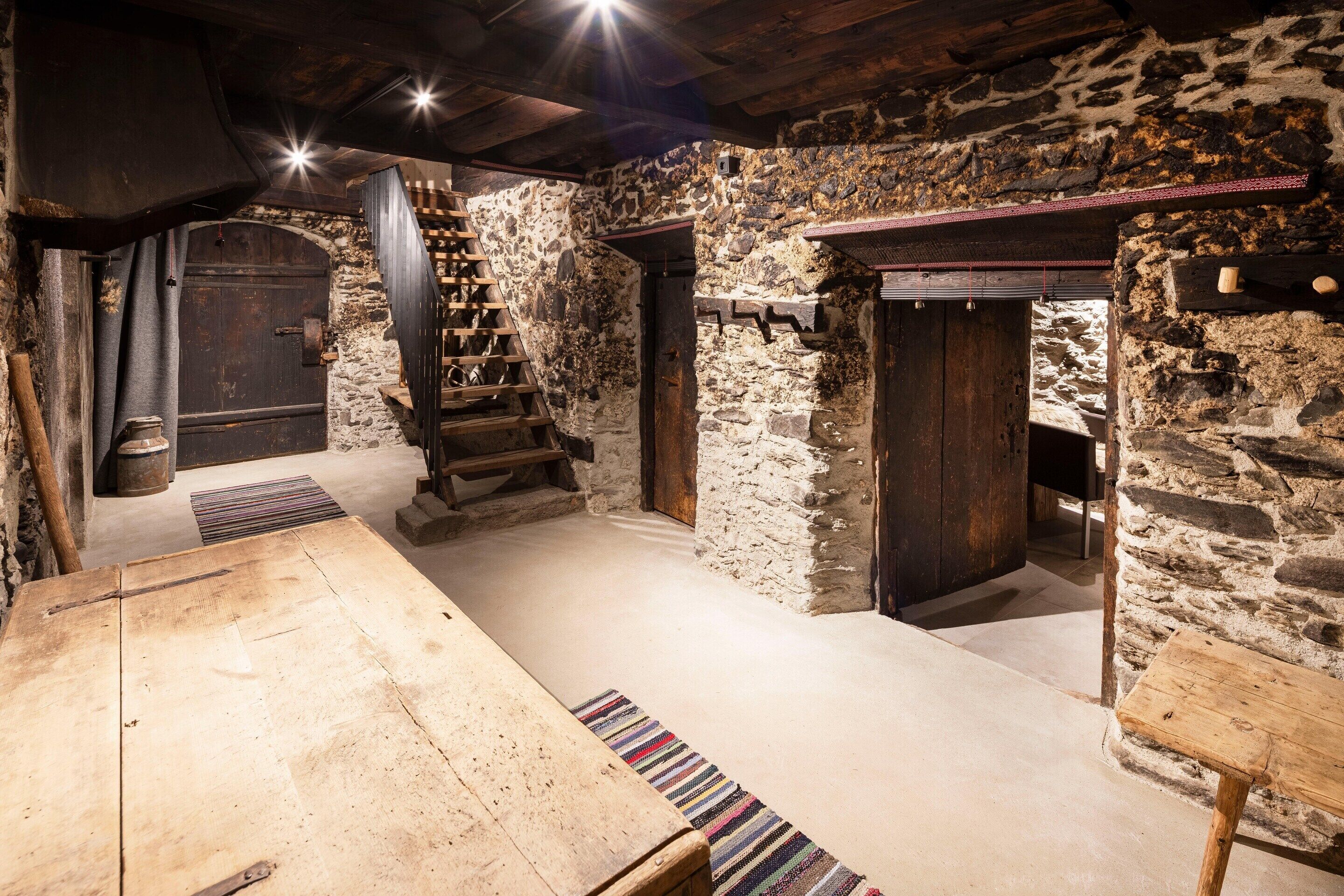 Interior rústico de piedra con una mesa de madera, una escalera y una chimenea.