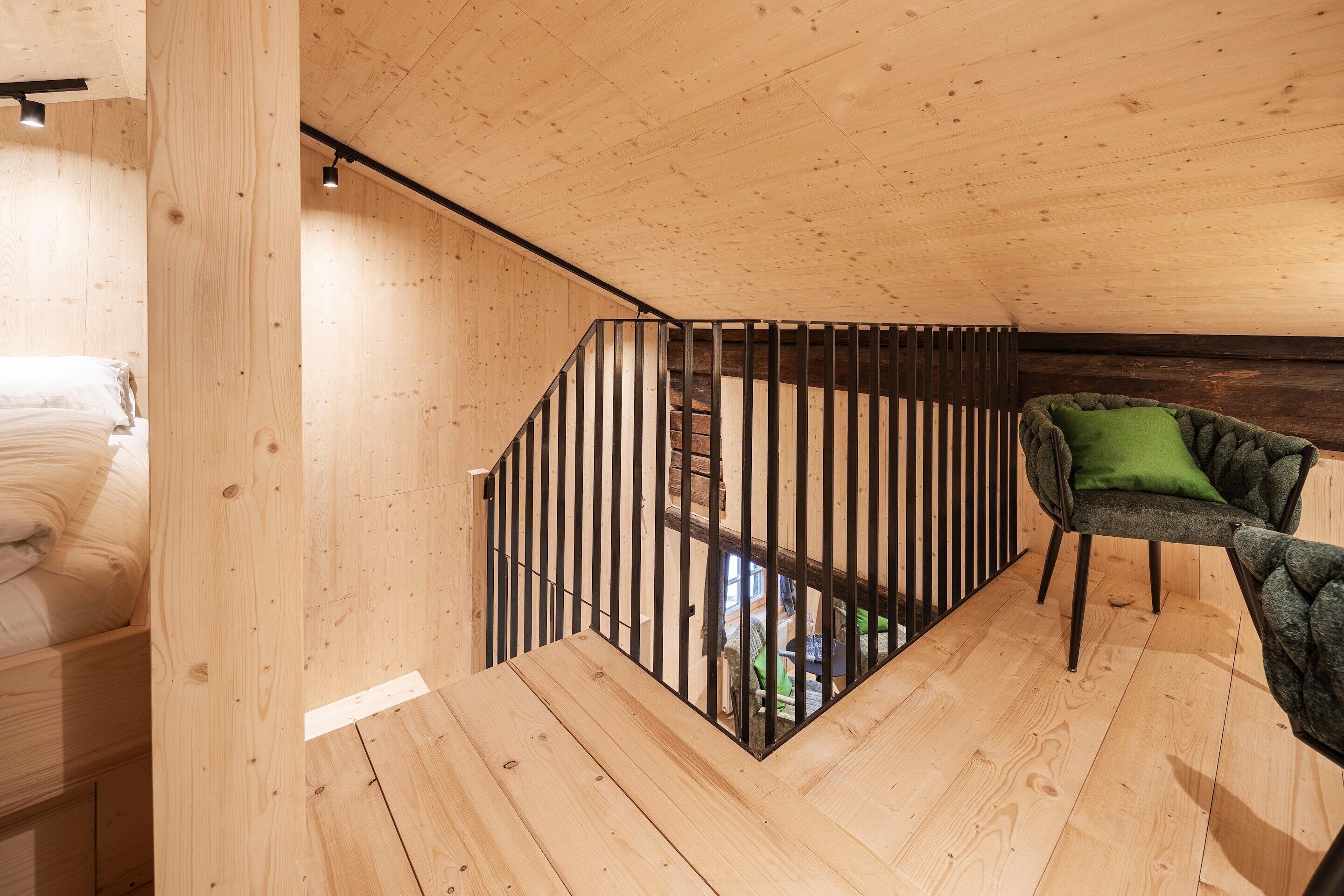 Un intérieur en bois avec un escalier, un fauteuil recouvert d’un tissu vert et un lit avec une literie blanche.