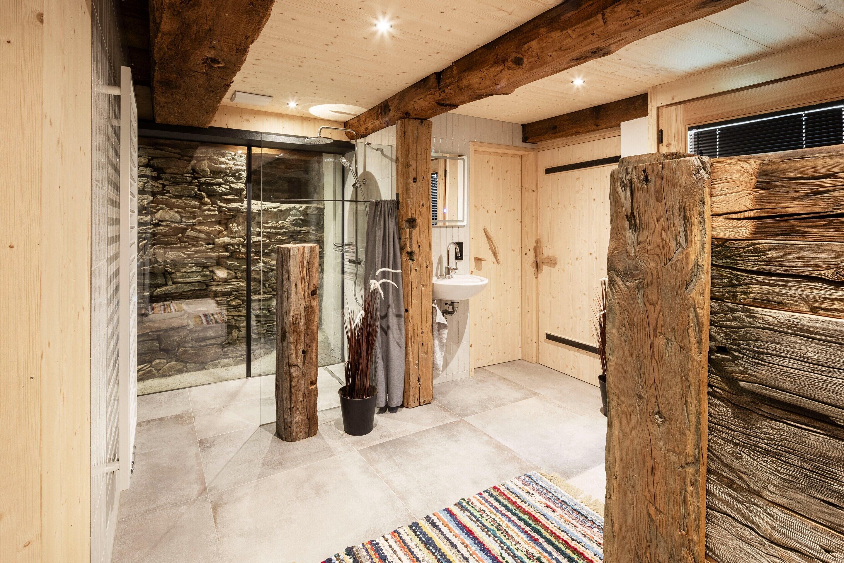 Ein Badezimmer mit Steinwand, Duschkabine aus Glas, Waschbecken und Holzbalken.