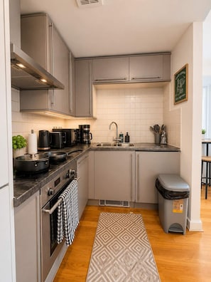 A modern kitchen with stainless steel appliances, a tiled backsplash, and a small trash bin. - Cardiff City Centre 2-Bed, 2-Bath Flat | Balcony | Coopers Court (Cardiff)