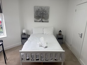 A bedroom with a bed, two bedside tables, a nightstand, a window, and a door. - Cozy Studio Flat - With Garden and Modern Amenities (Cardiff)
