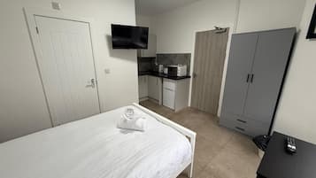 A bedroom with a bed, a TV mounted on the wall, a kitchenette with a microwave and toaster, and a grey wardrobe.