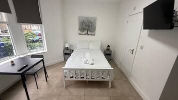 A bedroom with a bed, a desk, a chair, a nightstand, a lamp, a painting, and a TV mounted on the wall.