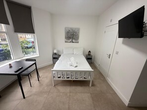 A bedroom with a bed, a desk, a chair, a nightstand, a lamp, a painting, and a TV mounted on the wall. - Cozy Studio Flat - With Garden and Modern Amenities (Cardiff)