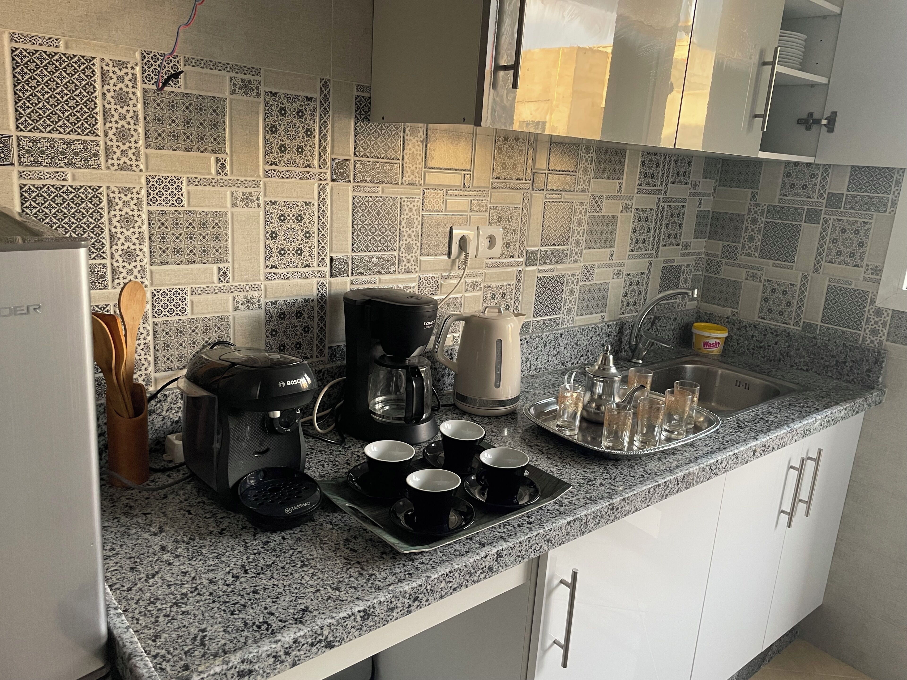 A kitchen with a patterned backsplash, a coffee maker, cups, and a tray with glasses.