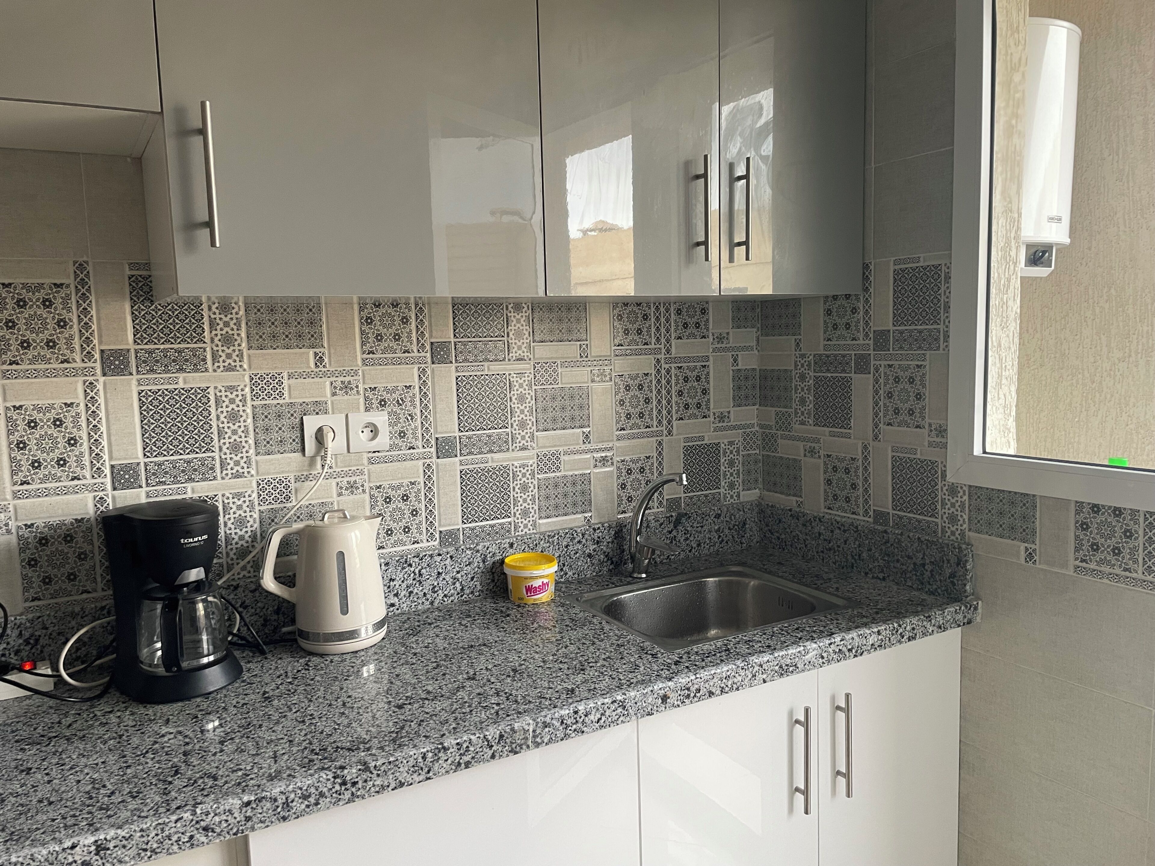 A kitchen with white cabinets, a granite countertop, a coffee maker, a kettle, and a sink.