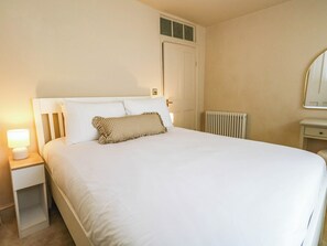 A neatly made bed with white linens and a checkered pillow, a bedside lamp, a closed door, and a radiator. - 123 On The Sea - The Cubby (Weymouth)