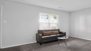 Ein Wohnzimmer mit grauem Sofa, einem kleinen weißen Tisch und einem Fenster mit Rollläden.