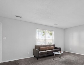 Ein Wohnzimmer mit grauem Sofa, einem kleinen weißen Tisch und einem Fenster mit Rollläden.
