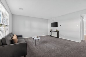 A modern living room with a grey carpet, a black sofa with cushions, a small white table, and a black entertainment unit with a TV. - Comfortable 2BR Retreat at Country Lakes (Indianapolis)