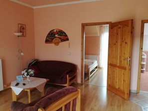A room with a wooden floor, a sofa, a table with a vase of flowers, a bed, and a wooden door. - Alsohegyi apartmanok by Interhome (Zalakaros-Thermal)