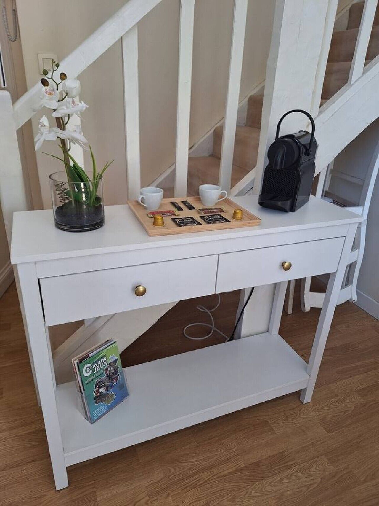 A white console table with a small shelf, a potted orchid, two white cups, a wooden tray with items, and a black bag.