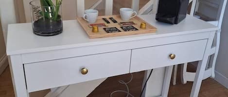 Una consola blanca con un estante pequeño, una orquídea en maceta, dos tazas blancas, una bandeja de madera con objetos y un bolso negro.