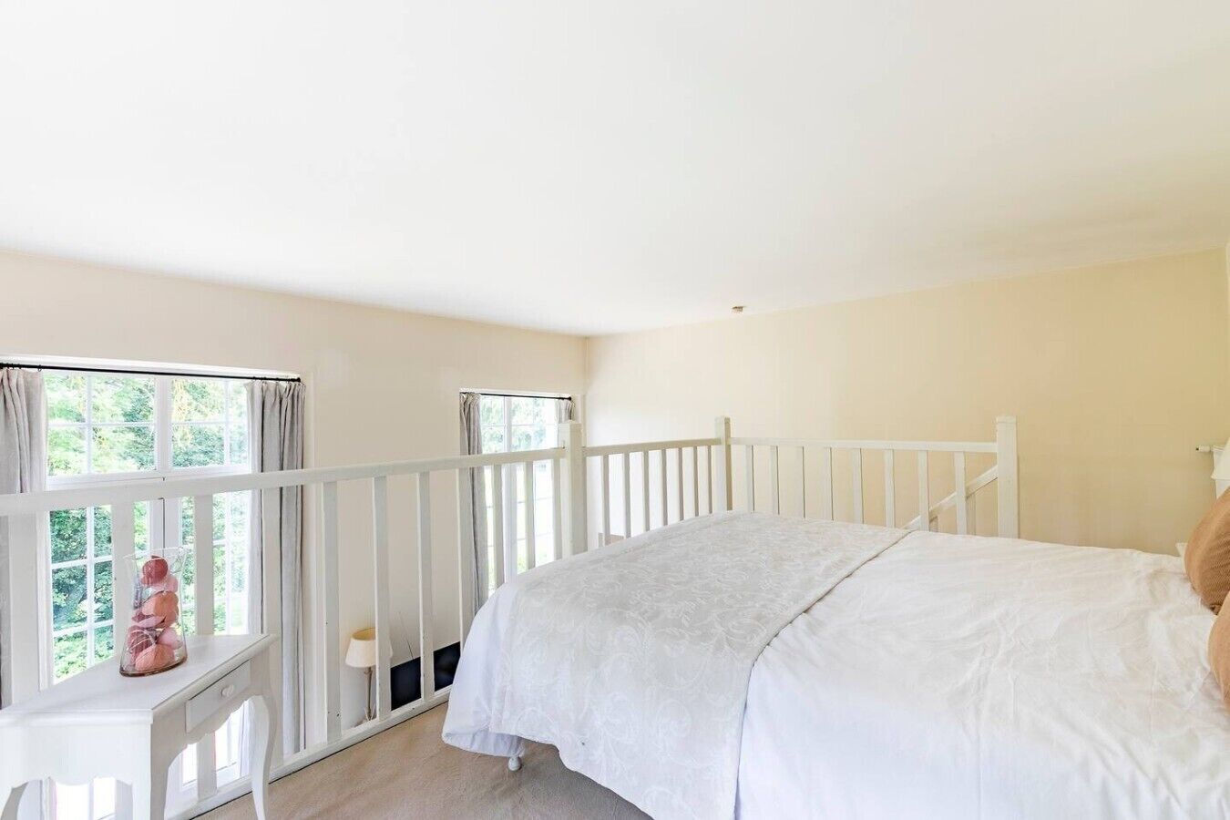 Quarto com uma cama de solteiro, janela com cortinas, uma mesinha com abajur e um corrimão branco.