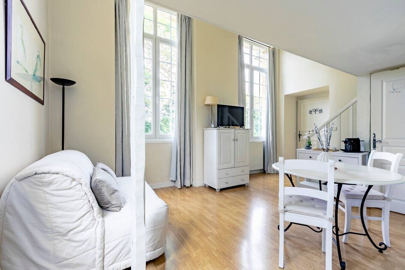 Un salon moderne avec un canapé blanc, une table à manger avec des chaises, un téléviseur posé sur un meuble et de grandes fenêtres avec des rideaux.