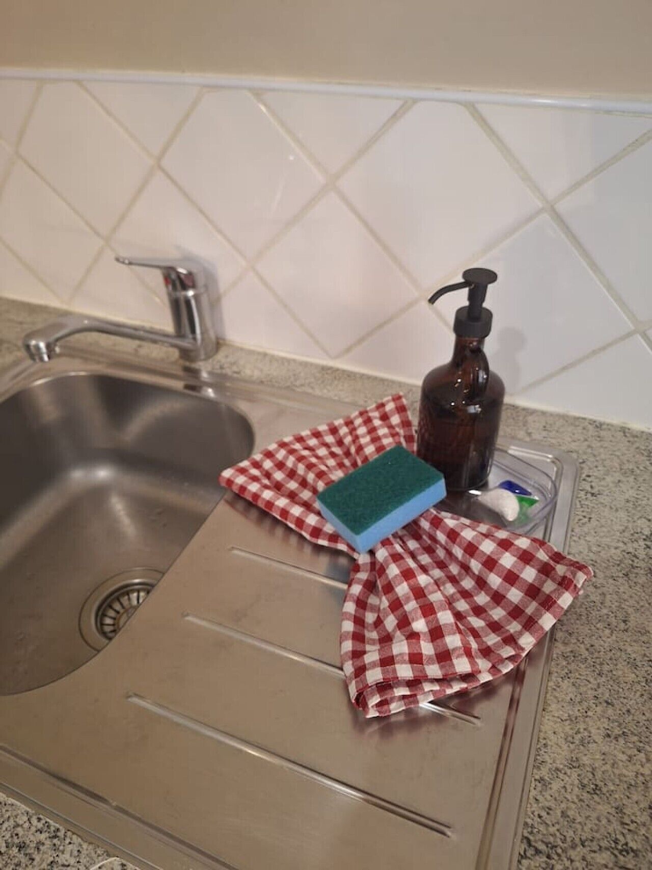 A kitchen sink with a faucet, a soap dispenser, a sponge, and a red and white checkered cloth.