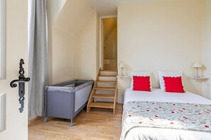 A bedroom with a bed, two bedside lamps, a small table, a grey storage unit, a wooden ladder, and a door with an ornate handle.