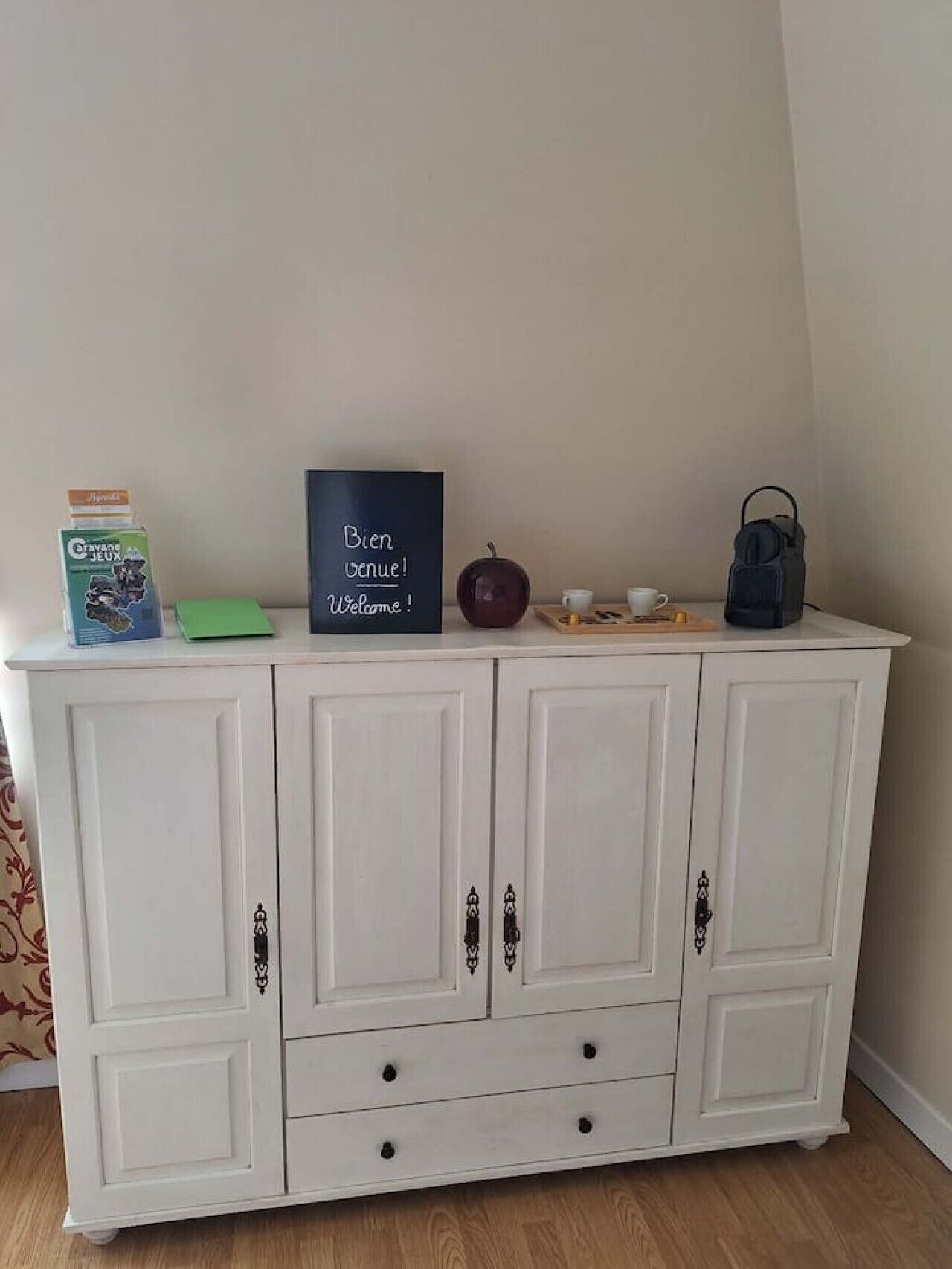Ein weißer Schrank mit Schubladen und Türen, eine Schiefertafel mit der Aufschrift 'Bien Venue!', ein roter Apfel, eine blaue Tasche und eine grüne Kiste.