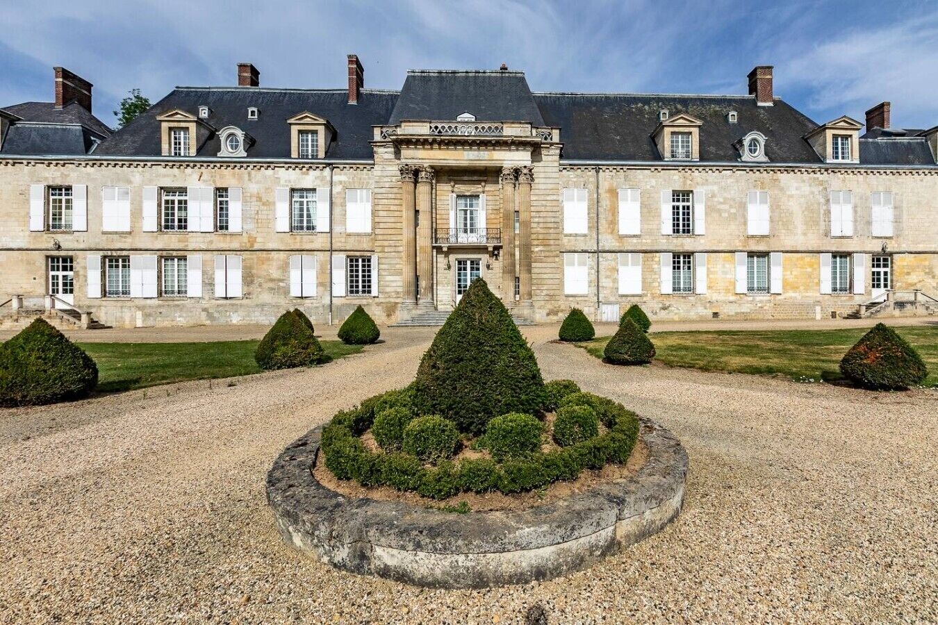 A large, historic building with a symmetrical facade, a central entrance, and a formal garden with trimmed hedges.