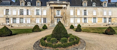 A large, historic building with a symmetrical facade, a central entrance, and a formal garden with trimmed hedges.