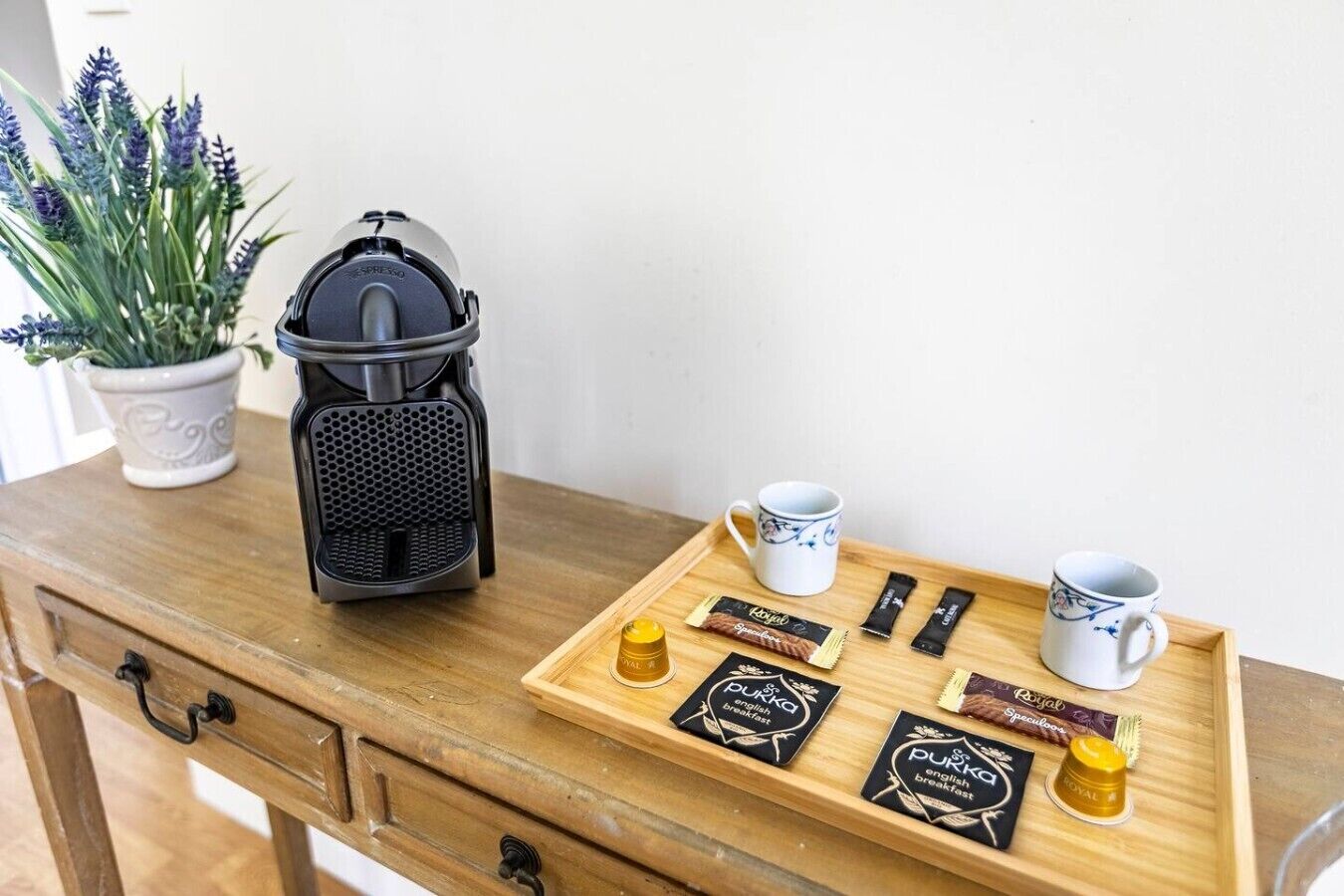 Una mesa de madera con una cafetera, dos tazas blancas, una bandeja con alimentos para el desayuno y una planta en maceta.