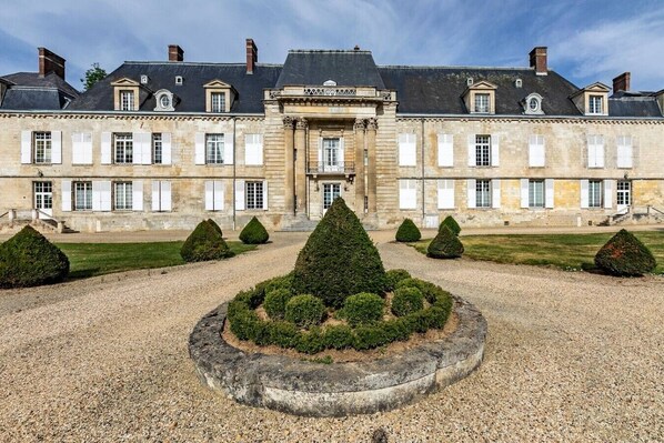 A large, historic building with a symmetrical facade, a central entrance, and a formal garden with trimmed hedges.