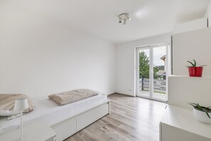 Ein schlicht eingerichtetes Schlafzimmer mit Bett, Nachttisch und Blick durch eine Schiebetür auf einen Balkon.