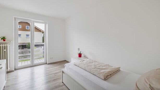 Une chambre à coucher minimaliste, avec un lit, une petite table de chevet, une plante rouge et une vue sur un espace extérieur accessible par une porte coulissante.