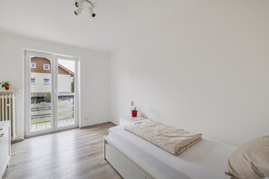 Ein schlichter Schlafbereich mit einem Bett, einem kleinen Nachttisch, einer roten Pflanze und Blick durch eine Schiebetür auf einen Außenbereich.