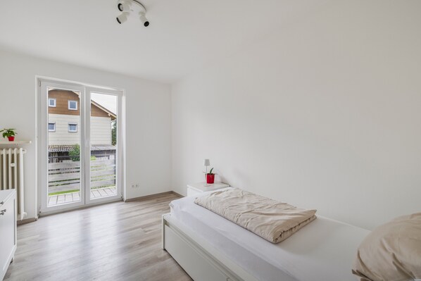 A minimalist bedroom with a bed, a small bedside table, a red plant, and a view through a sliding door to an outdoor area.