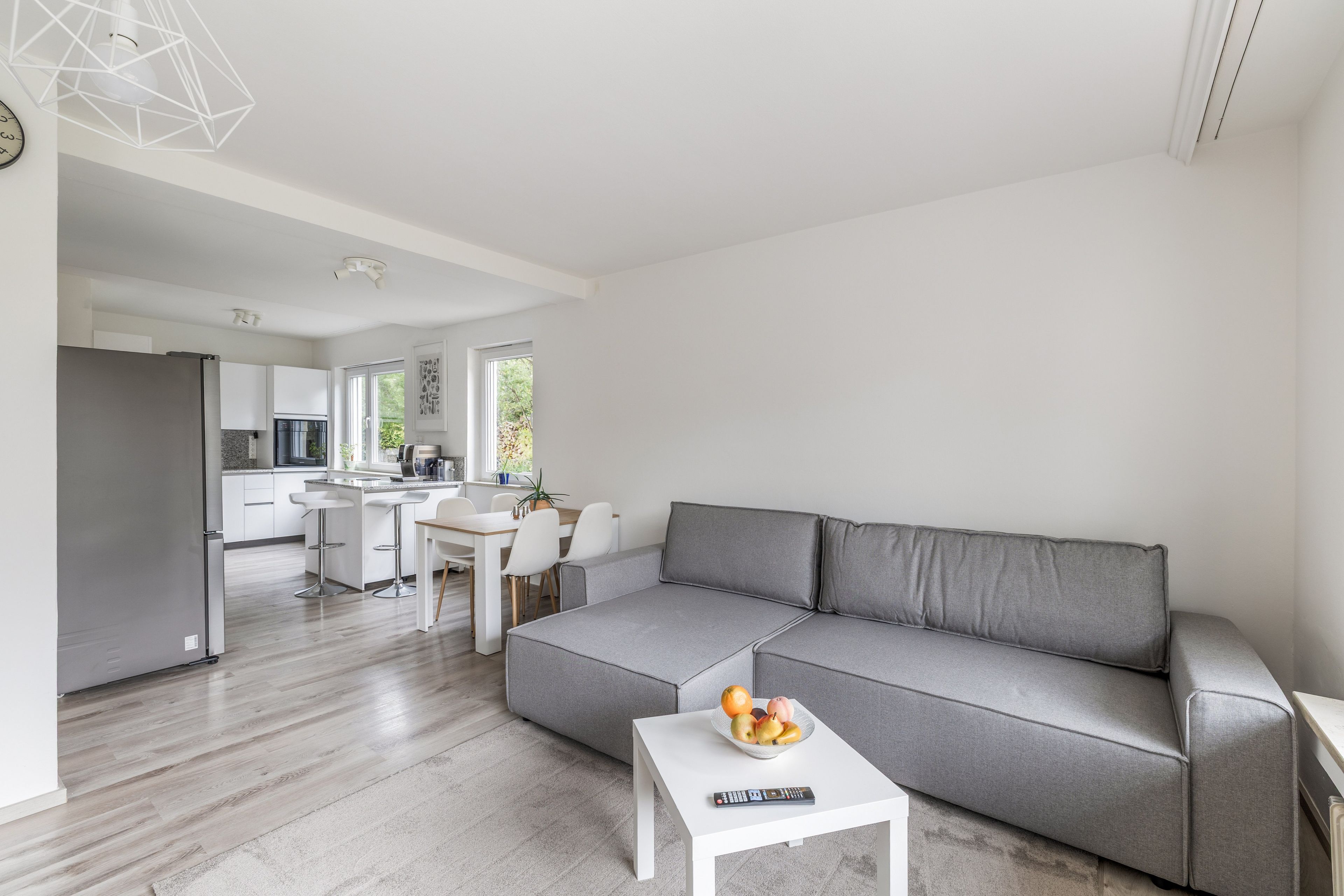 Un salon moderne avec un canapé gris, une petite table blanche sur laquelle se trouvent des fruits et une télécommande, et une cuisine en arrière-plan.