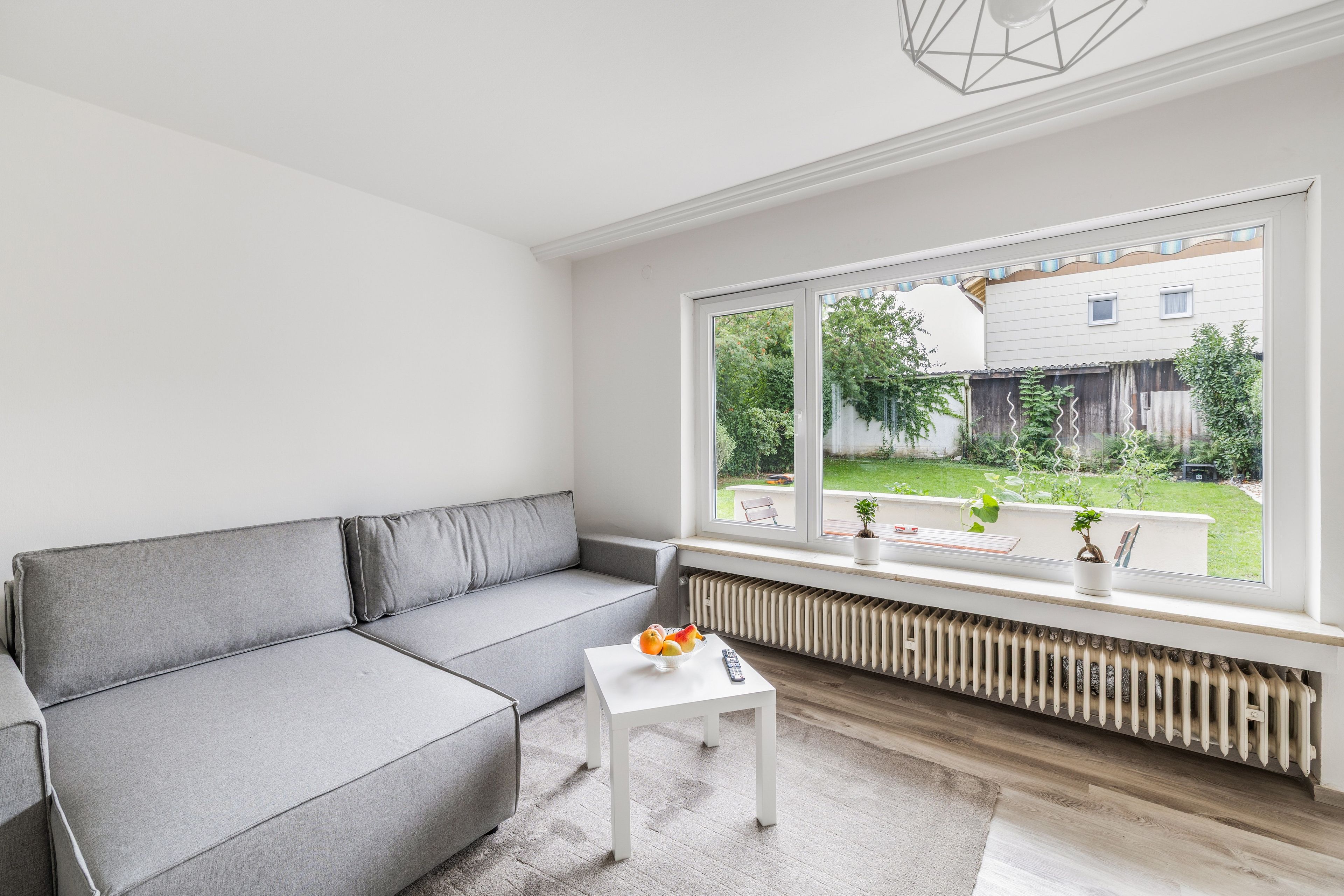 Ein modernes Wohnzimmer mit einem grauen Sofa, einem kleinen weißen Tisch und einem großen Fenster mit Blick auf einen Garten und ein Haus.