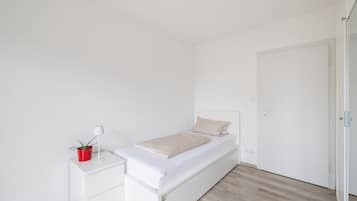 Ein schlichter Schlafzimmerbereich mit einem Einzelbett, einem Nachttisch mit Lampe und einer Topfpflanze sowie einer Tür.