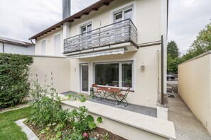 A two-story house with a balcony, a dining area with a table and chairs, and a garden with plants. - Holiday Home 'AB12' with Private Terrace, Garden and Wi-Fi (Oberhaching)