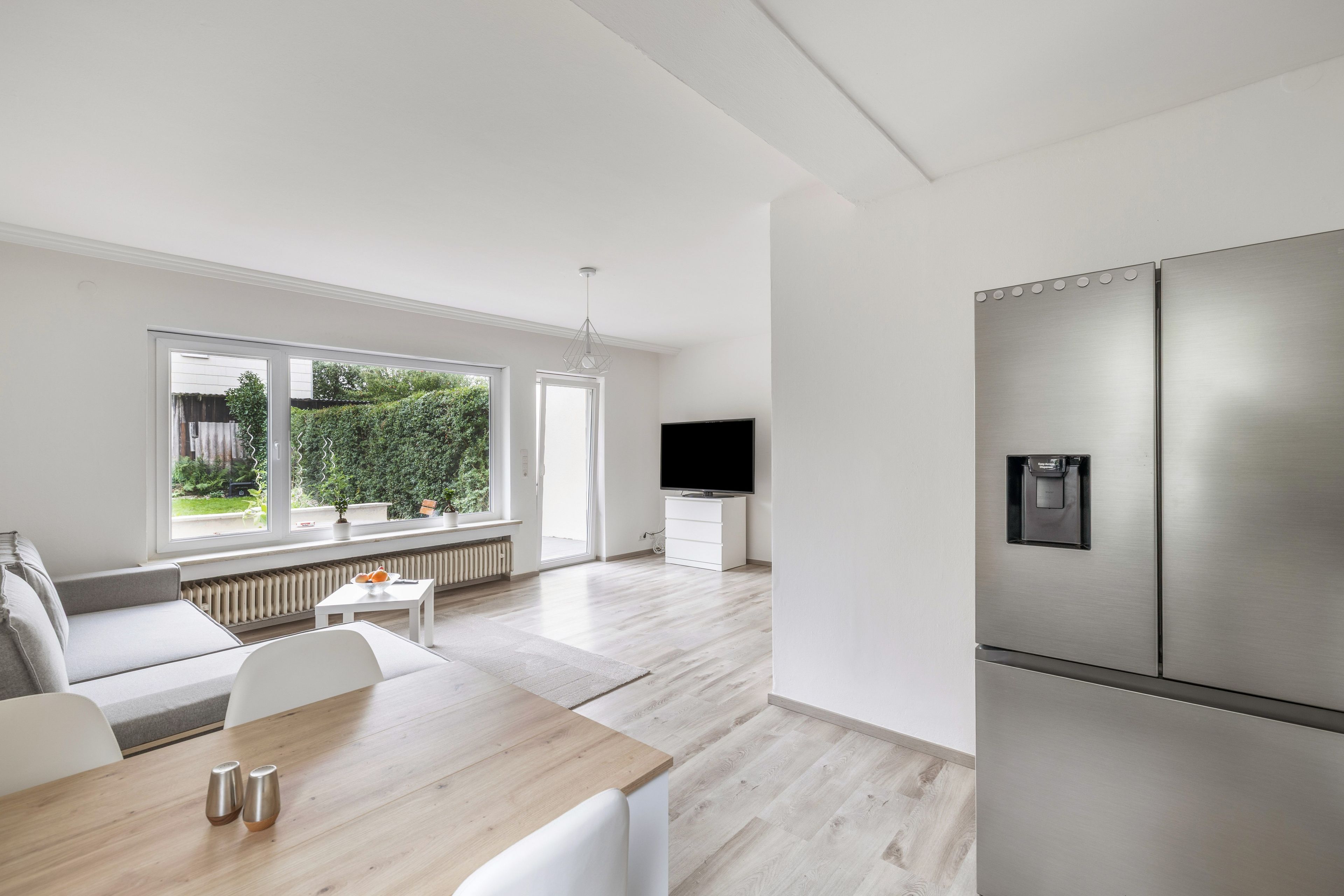 A modern kitchen with a stainless steel refrigerator, a dining area with a wooden table, and a large window with a view of greenery.