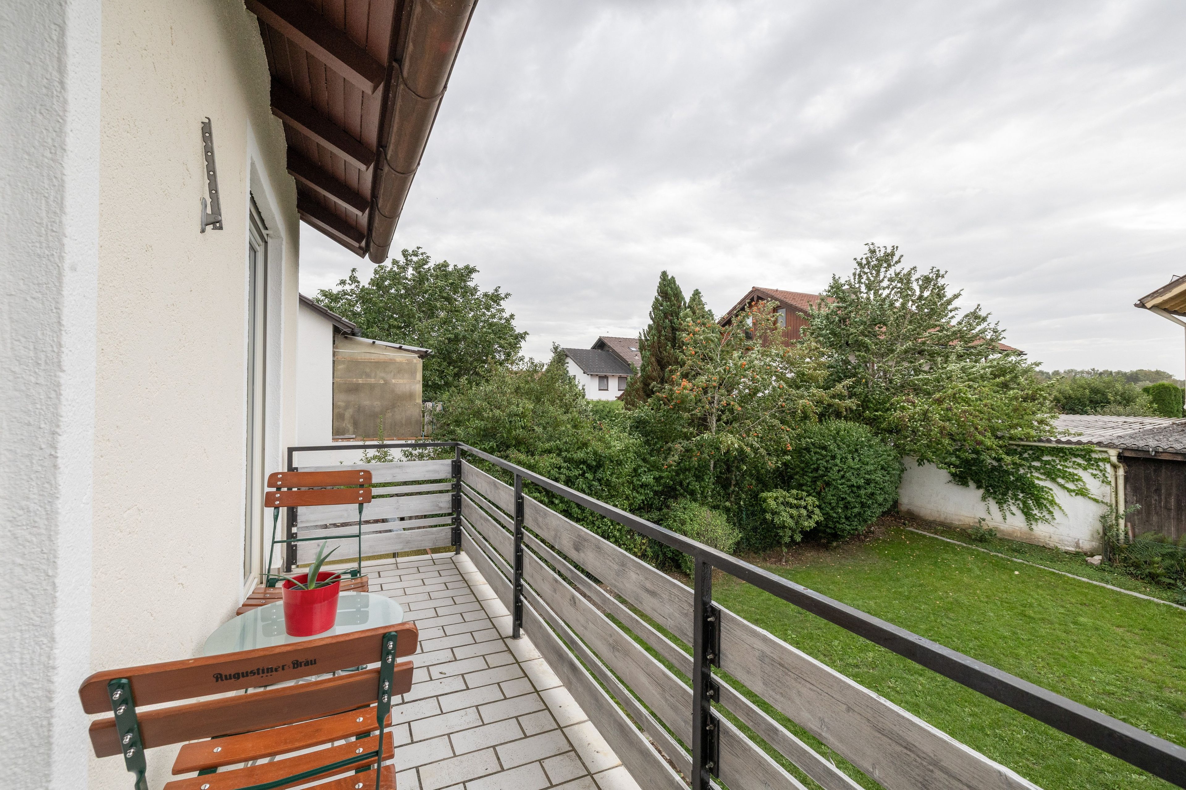 A balcony with a table and chairs, a potted plant, and a view of a green garden and buildings.