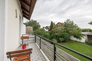 A balcony with a table and chairs, a potted plant, and a view of a green garden and buildings.