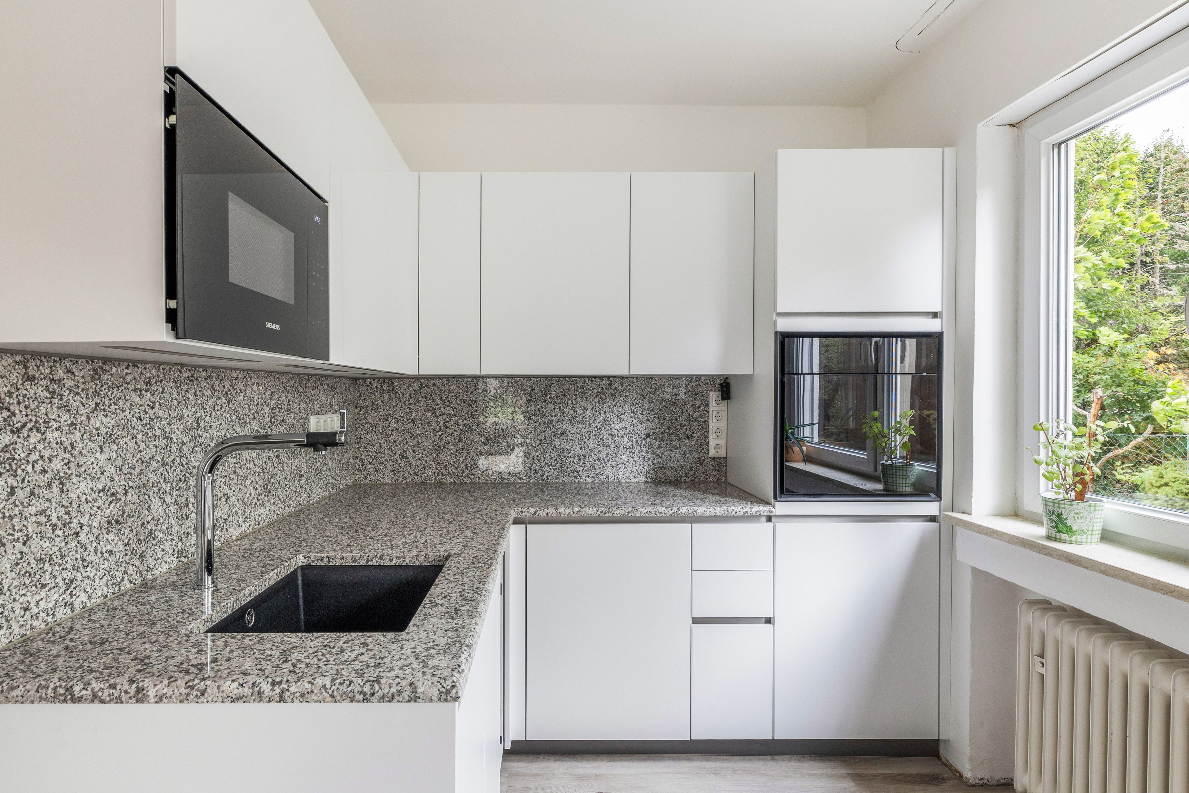 A modern kitchen with granite countertops, white cabinetry, a built-in microwave, and a window with a view of greenery.