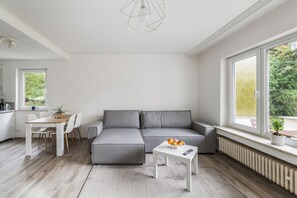 A modern living room with a grey sectional sofa, a dining table with chairs, and a small white coffee table with fruit. - Holiday Home 'AB12' with Private Terrace, Garden and Wi-Fi (Oberhaching)
